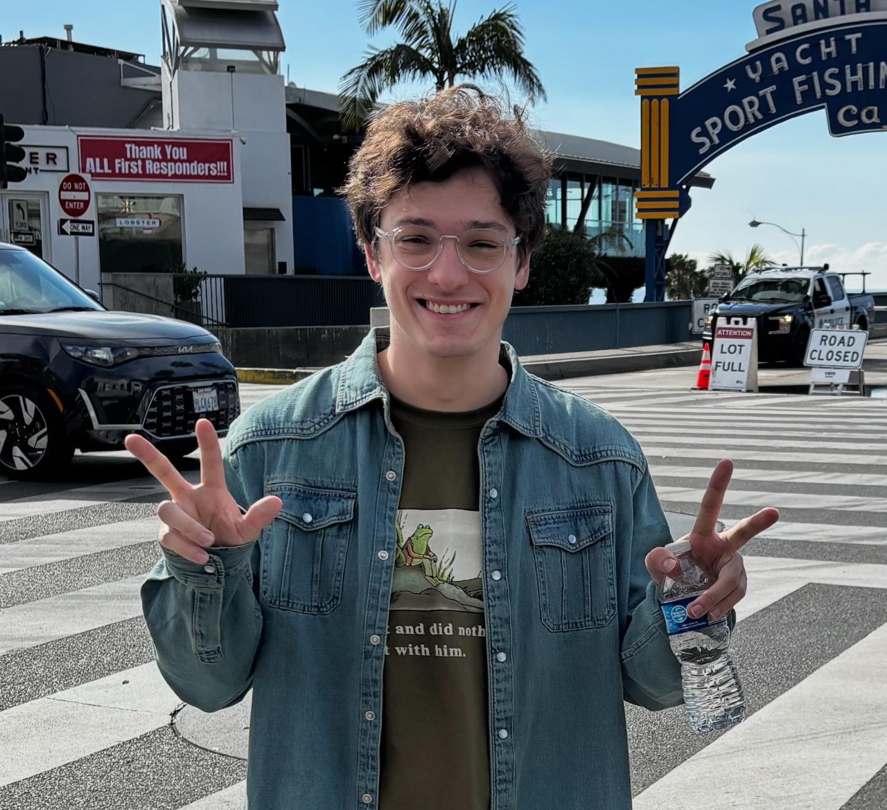 Foto de Enrico Chiosini sorrindo, vestindo uma camisa azul clara e um casaco escuro, com um fundo desfocado de árvores e céu azul.
